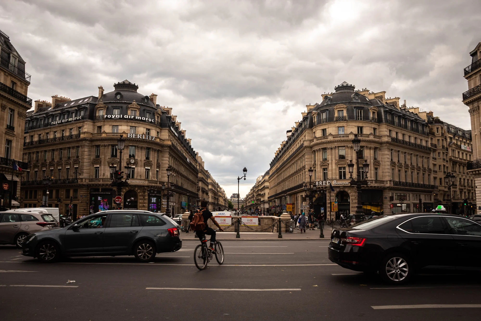 Ciclista attraversa un incrocio parigino tra le auto, palazzi haussmanniani sotto un cielo nuvoloso