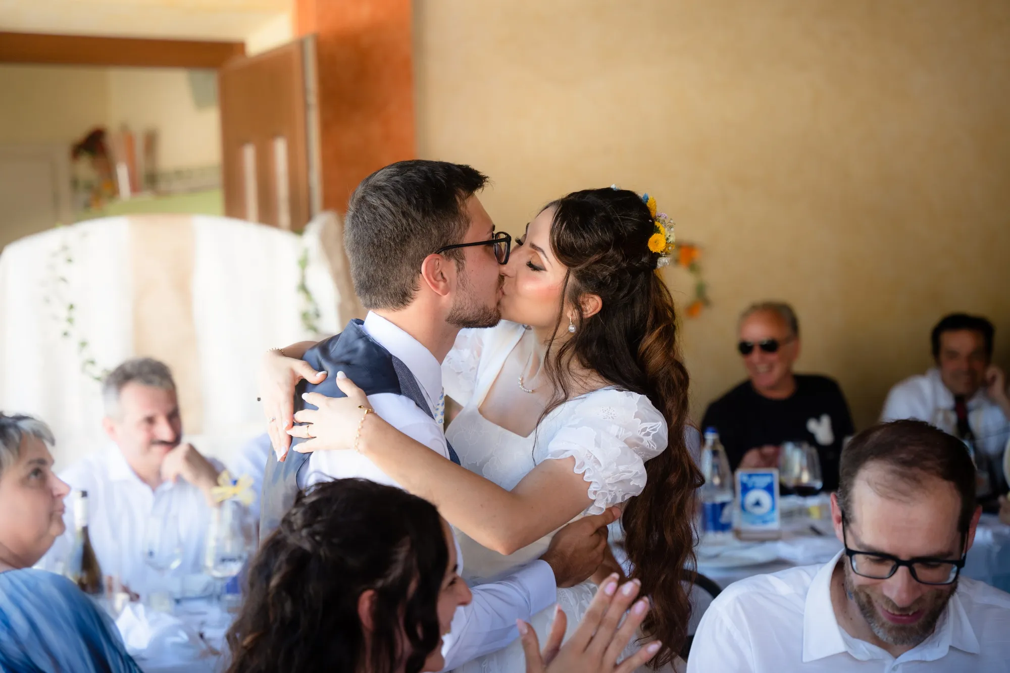 Fra i tavoli del pranzo gli sposi si baciano abbracciati, gli invitati applaudono intorno