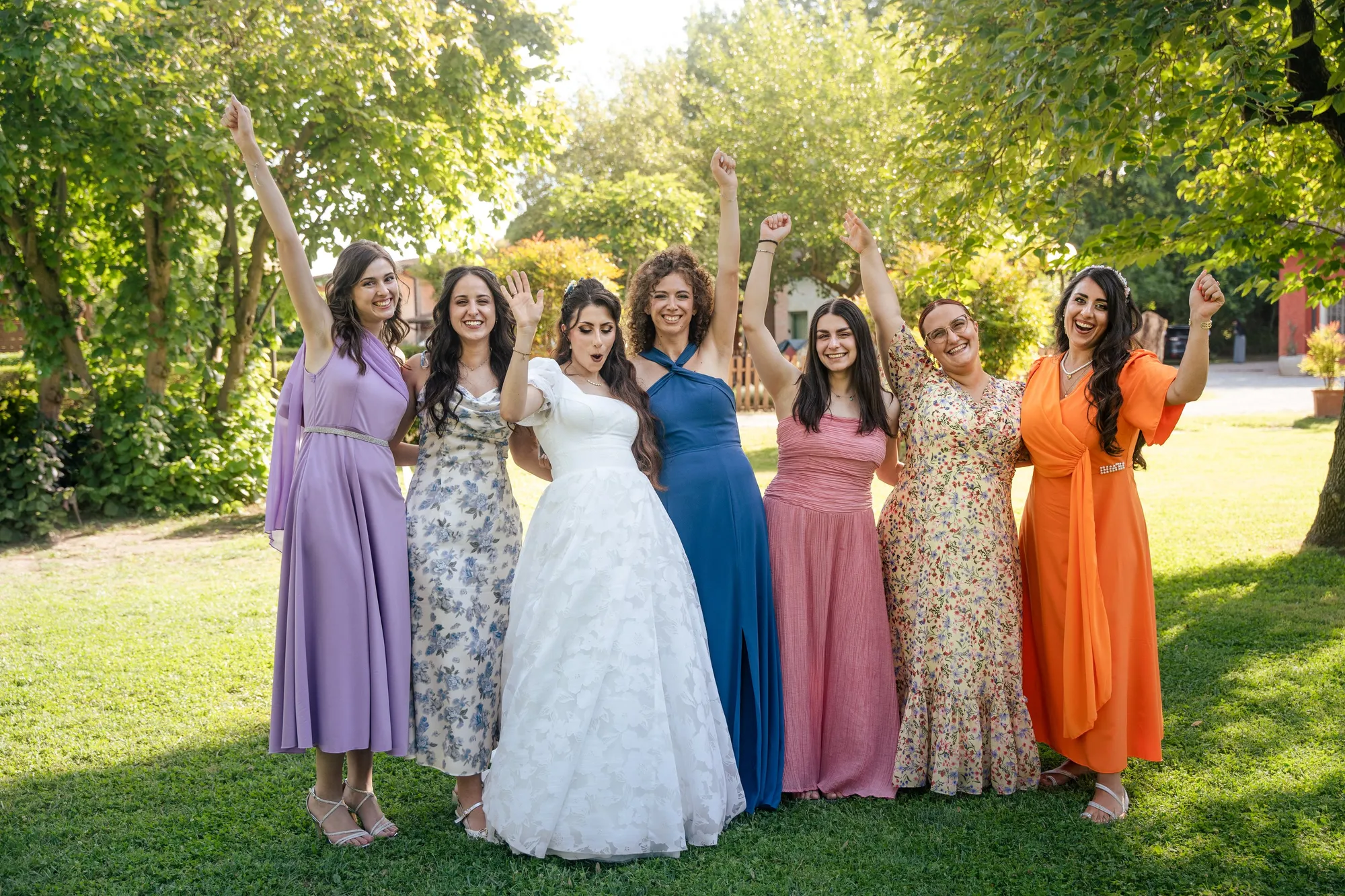 La sposa e sei damigelle alzano le braccia esultando sul prato del giardino