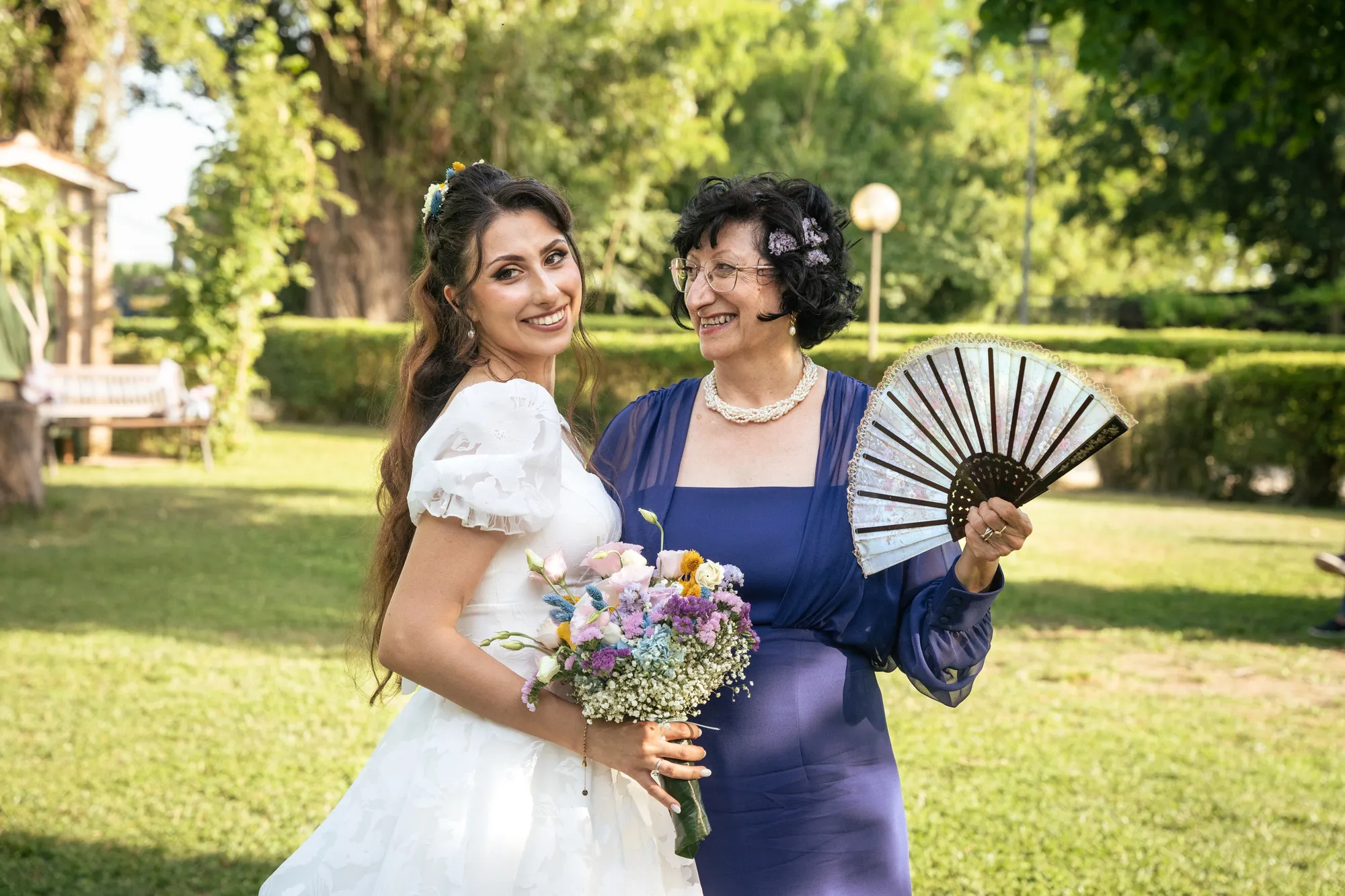 La sposa ride accanto a un'invitata in abito viola che si fa aria con un ventaglio nero