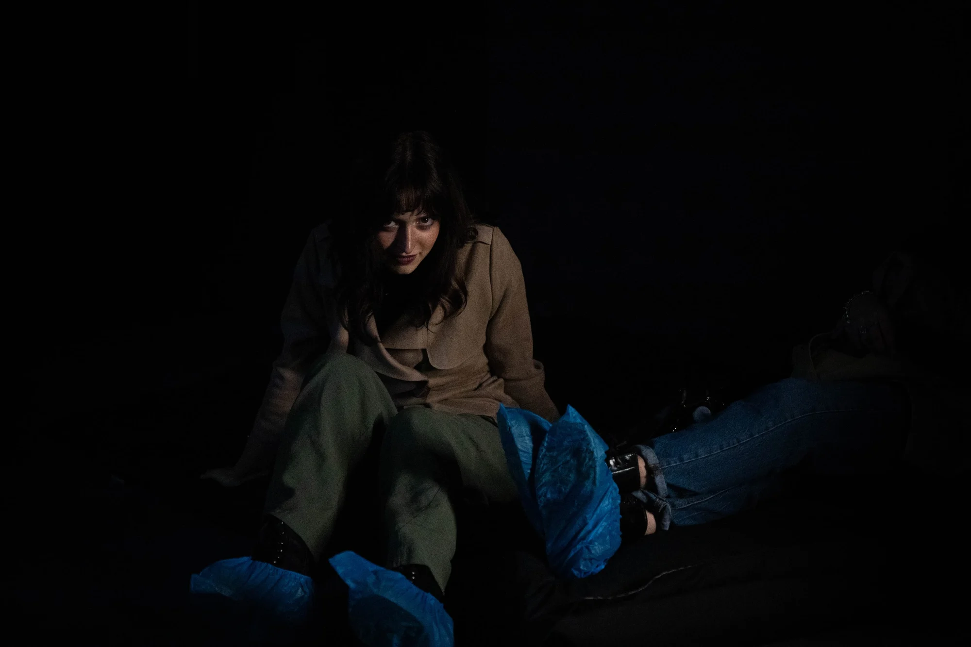 Ragazza con cappotto beige seduta nel buio, copriscarpe blu in primo piano, sguardo rivolto all'obiettivo